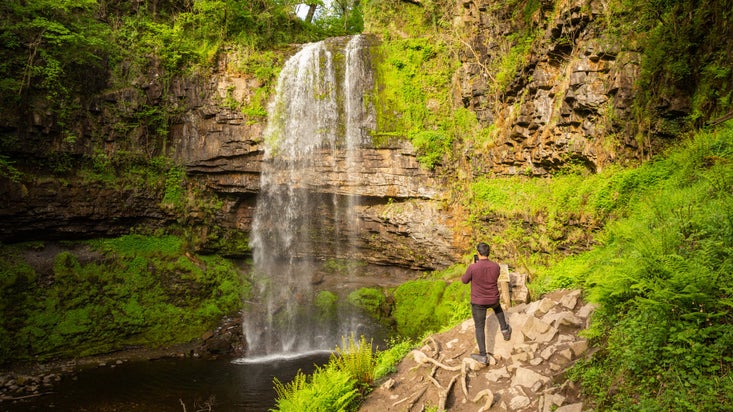 Visitor taking picture of waterfall with his phone while standing on small cliff above pool of water at Henrhyd Falls in the Brecon Beacons, Wales
